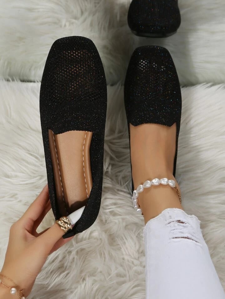 Svarta glittrande slip-on-skor i mesh sedda uppifrån, den ena hållen i handen så att den mjuka läderliknande innersulan syns, på en vit lurvig matta. 1