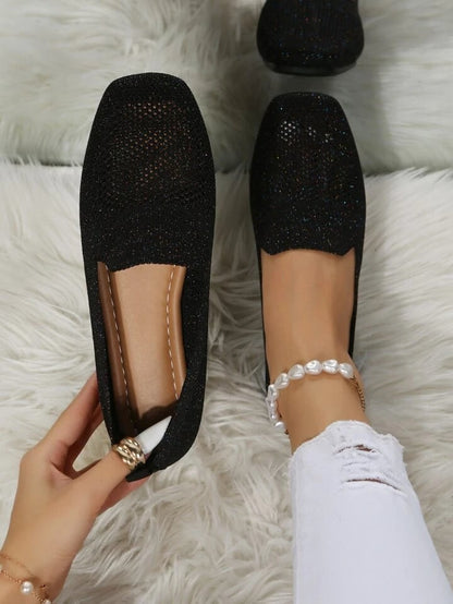 Svarta glittrande slip-on-skor i mesh sedda uppifrån, den ena hållen i handen så att den mjuka läderliknande innersulan syns, på en vit lurvig matta. 1