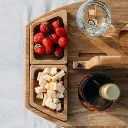 Holz-Picknicktisch mit Erdbeeren, Käsewürfeln, Weinglas und Flasche auf hellem Hintergrund.