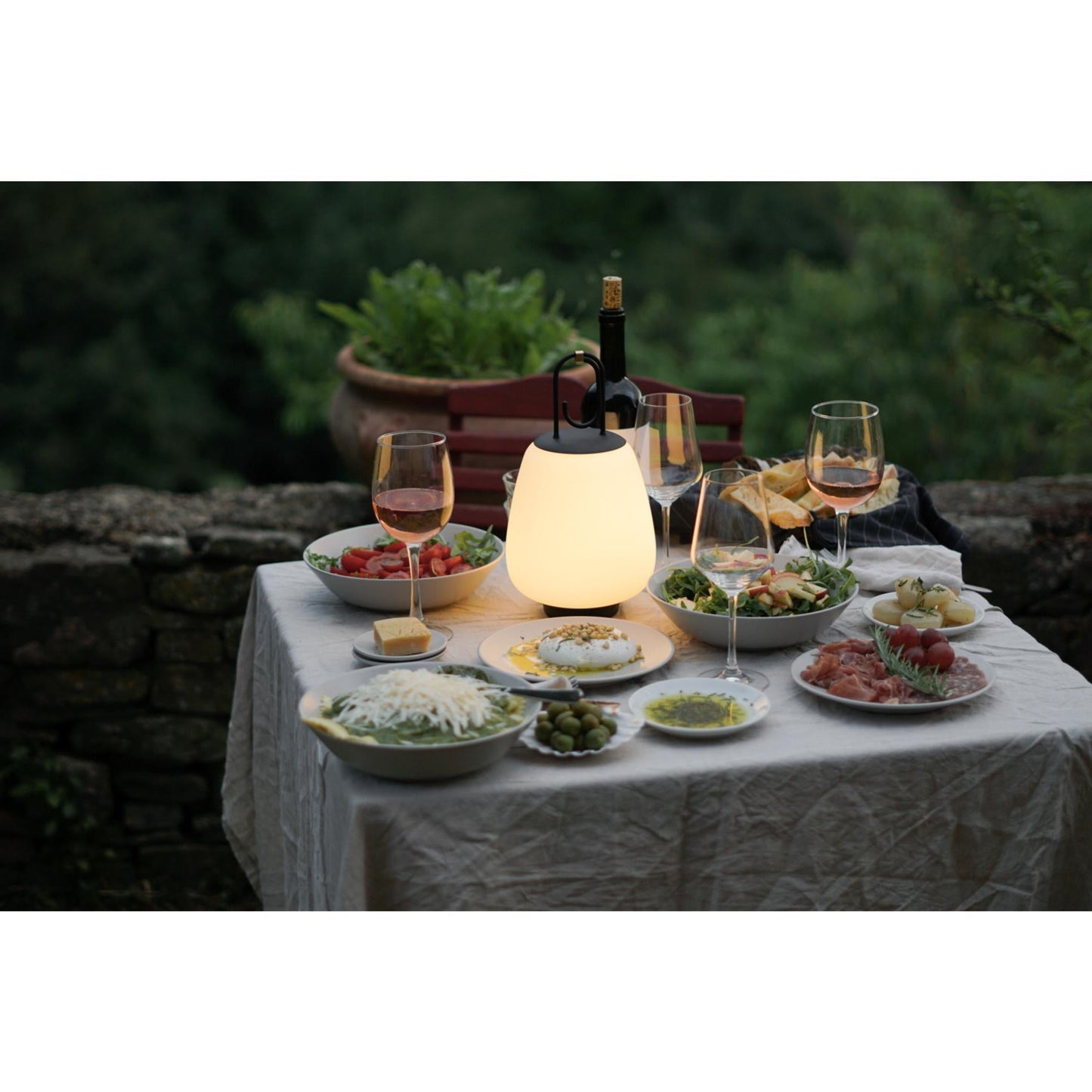 Tisch mit Speisen, Weingläsern und leuchtender Lampe im Freien, Abendessen im Garten.