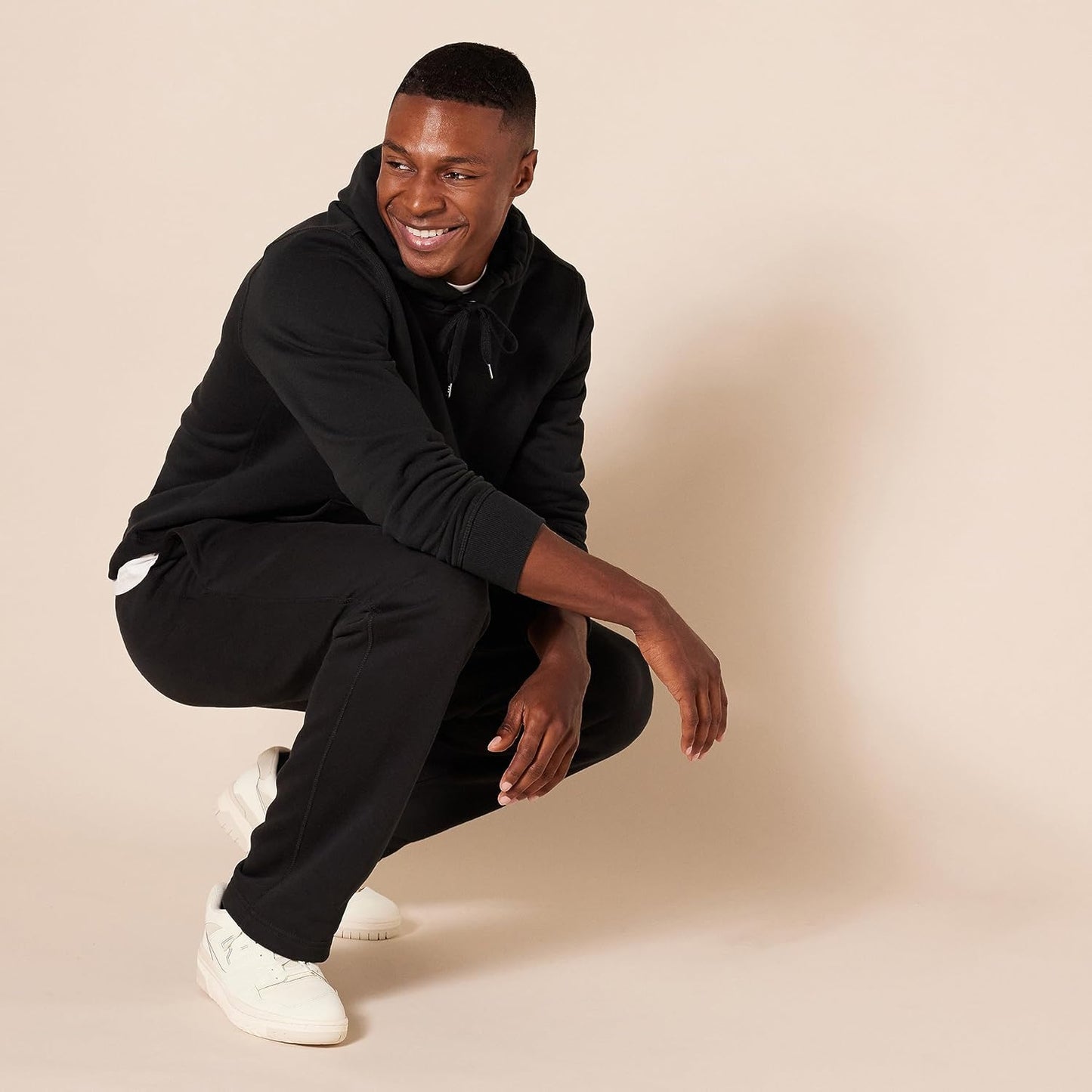 Mann in schwarzem Hoodie und Jogginghose, weiße Sneaker, lächelnd, beige Hintergrund.