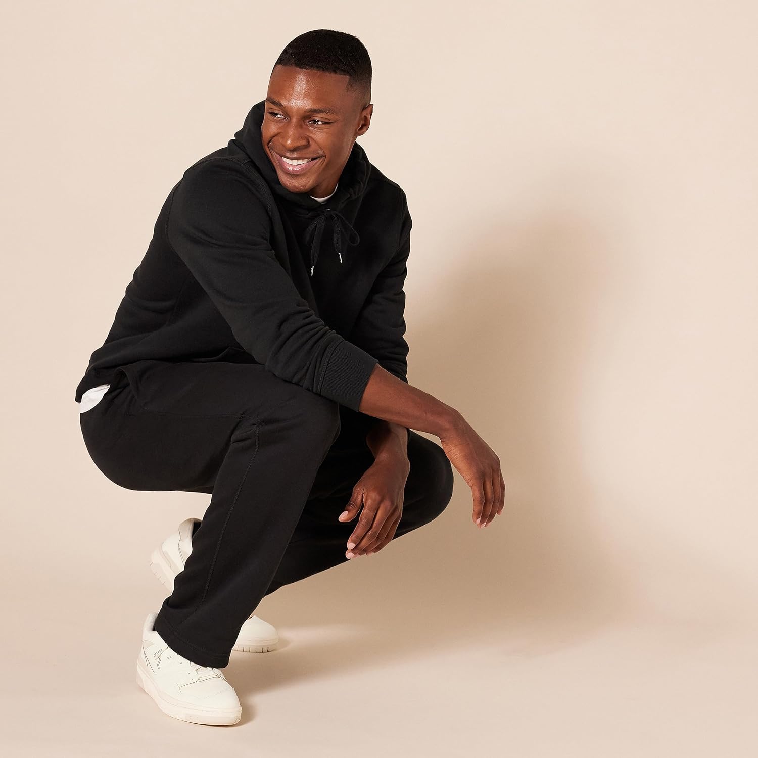 Mann in schwarzem Hoodie und Jogginghose, weiße Sneaker, lächelnd, beige Hintergrund.