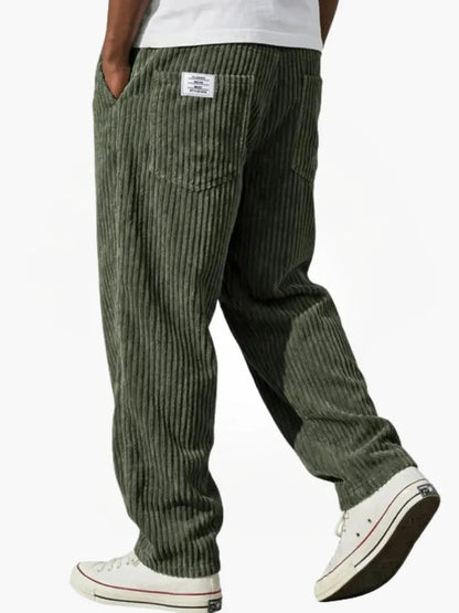 Olivgrön manchesterbyxa bakifrån på gående modell med vit t-shirt, bakficka, liten etikett och vita sneakers. 7