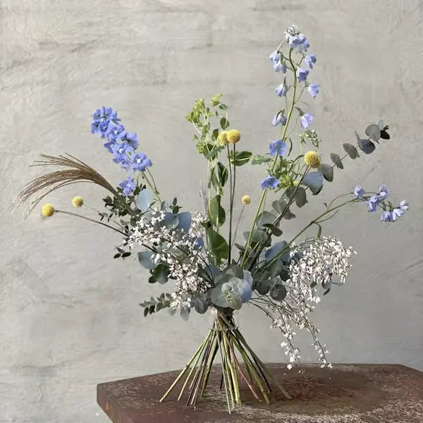 Fritt arrangerad bukett med blå blommor, gula bollar och eukalyptus, med stjälkarna samlade i en spiral mot en grå vägg. 17