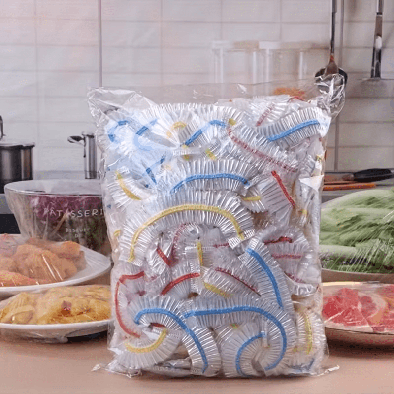 Stor genomskinlig påse full av elastiska engångslock med blå, röda och gula kanter, uppställd i ett kök bredvid övertäckta maträtter. 1