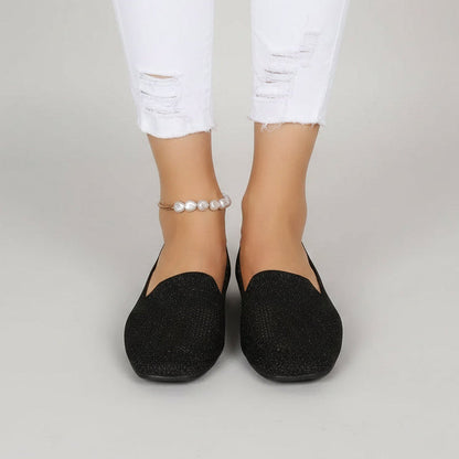 Svarta glittrande slip-on-skor med fyrkantig tå sedda framifrån på en modell i trasiga vita jeans och pärlankelkedja mot ljusgrå bakgrund. 2