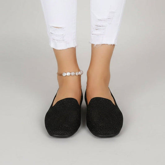 Svarta glittrande slip-on-skor med fyrkantig tå sedda framifrån på en modell i trasiga vita jeans och pärlankelkedja mot ljusgrå bakgrund. 2