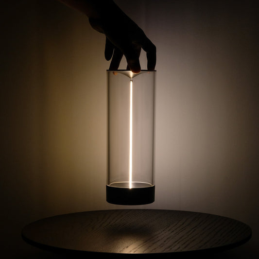 Hand hält moderne LED-Tischlampe, zylindrisch, transparent, auf Holztisch, minimalistisch.