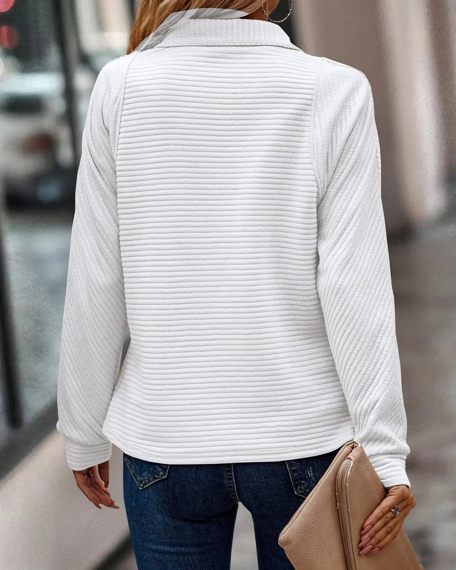 Frau in weißem Strickpullover und blauer Jeans, hält beige Clutch, Stadtstraße im Hintergrund.