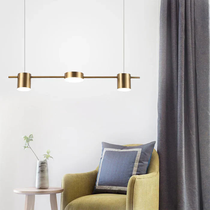 Moderne goldene Pendelleuchte über grünem Sessel, grauer Vorhang im Hintergrund.