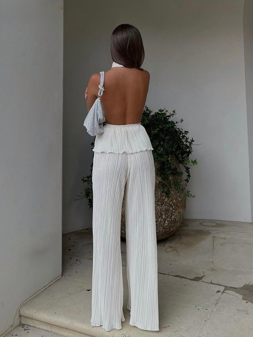 Frau in weißem, rückenfreiem Jumpsuit mit Plissee, graue Tasche, elegantes Sommeroutfit.