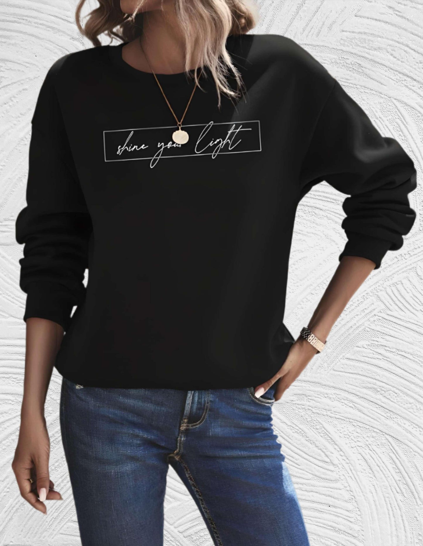 Frau in schwarzem Sweatshirt mit "shine your light" Aufdruck, blaue Jeans, lässiger Stil.