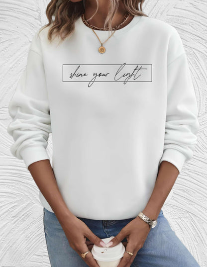 Frau in weißem Sweatshirt mit "shine your light" Aufdruck, Jeans, Kaffeetasse, Schmuck.