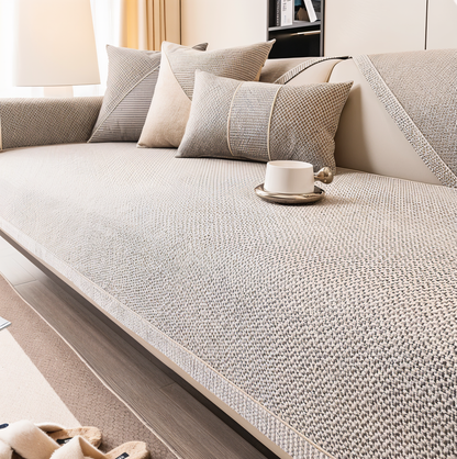 Soffa med grå-beige överdrag i grovt vävd struktur, kuddar och en vit kopp på sittytan. 1