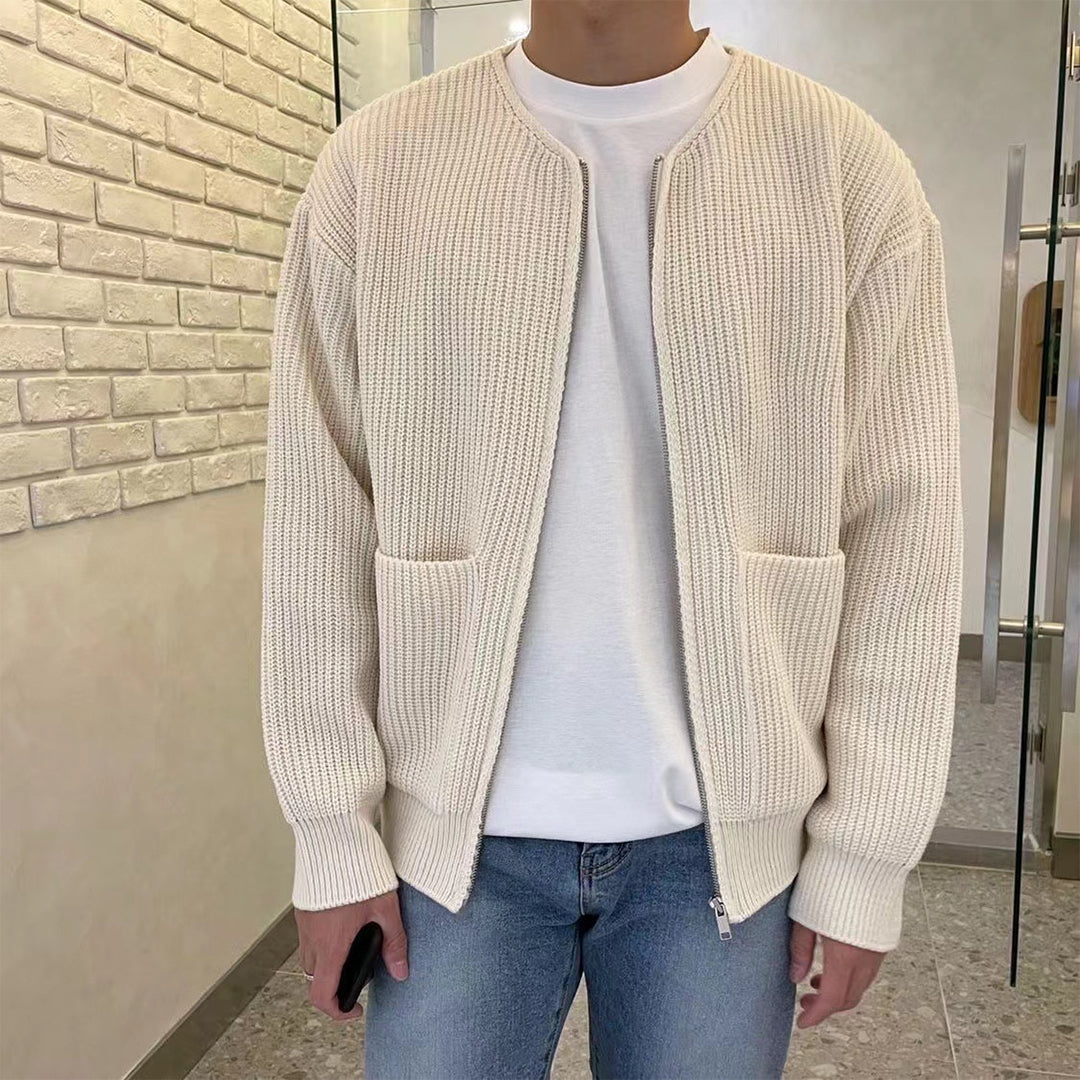 Mann in cremefarbener Strickjacke, weißem T-Shirt und blauer Jeans in modernem Innenraum.