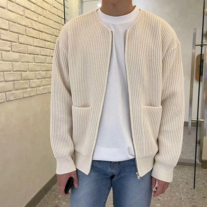 Mann in cremefarbener Strickjacke, weißem T-Shirt und blauer Jeans in modernem Innenraum.