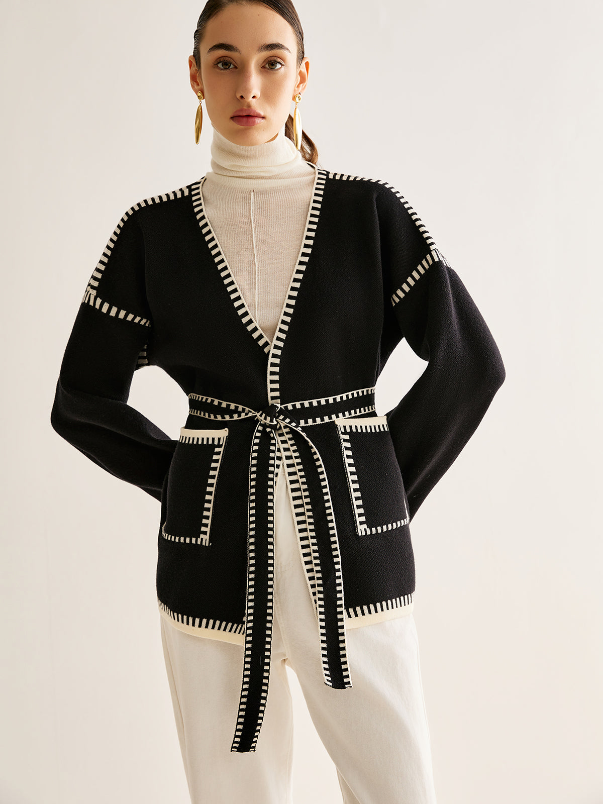 Frau in schwarzer Strickjacke mit Gürtel, weißem Rollkragenpullover und weißen Hosen.
