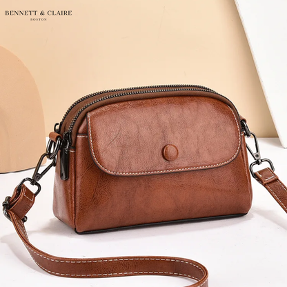 Crossbodyväska i konjaksbrunt skinnlook med framficka, dubbla blixtlås och vit kontrastsöm, fotograferad snett framifrån mot beige bakgrund. 1