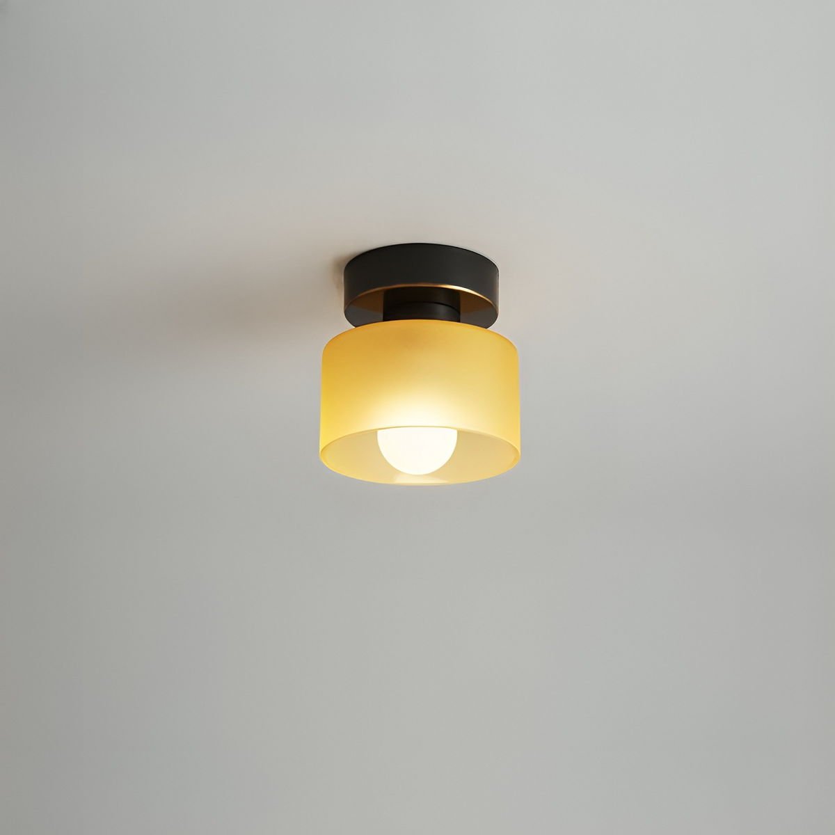 Takspotlight med svart fäste och gul frostad glasskärm, tänd glödlampa lyser varmt, mot grå bakgrund. 8