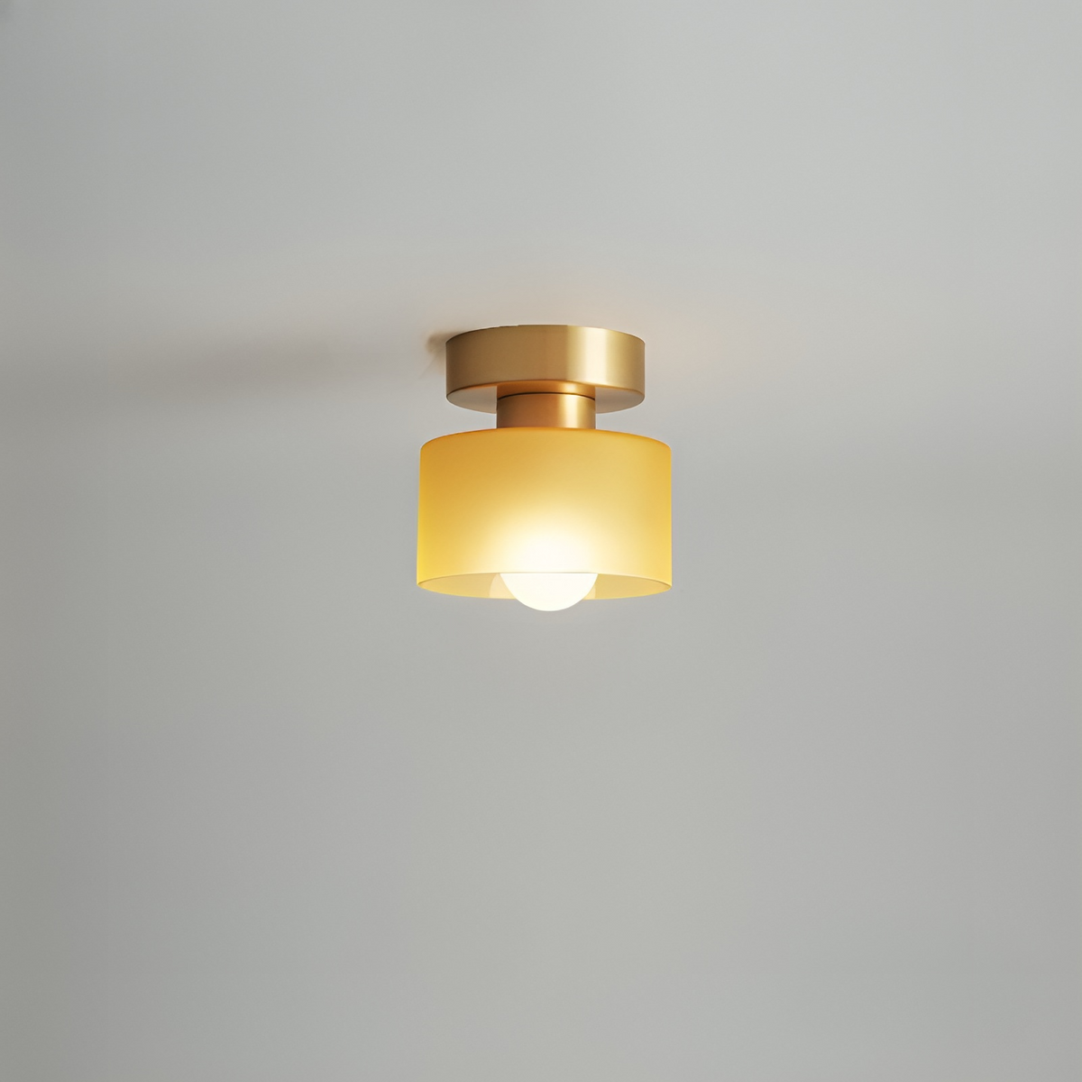 Takspotlight med guldfärgat fäste och gul frostad glasskärm, tänd glödlampa, mot ljusgrå bakgrund. 9