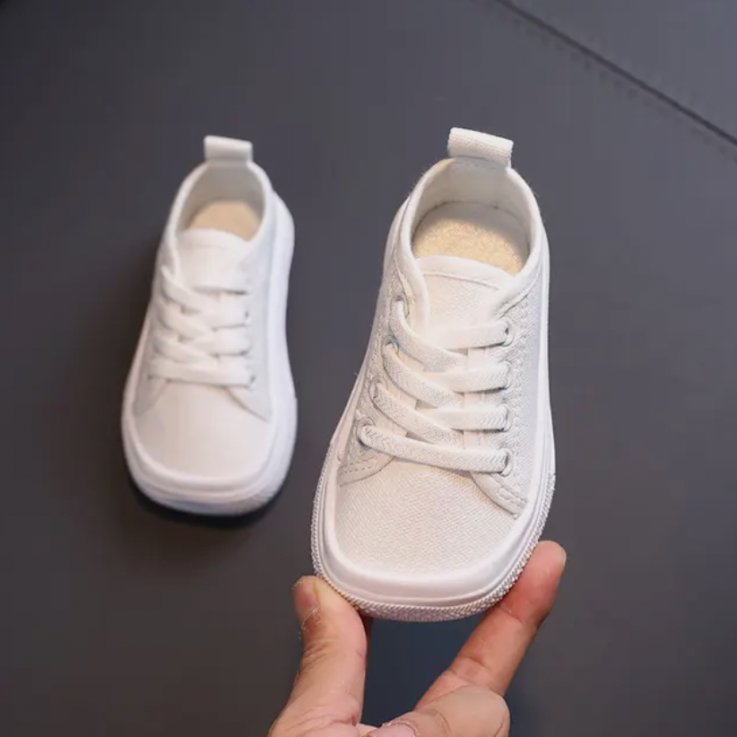 Weiße Kinderschuhe aus Canvas mit Schnürsenkeln, handgehalten, auf dunklem Hintergrund.