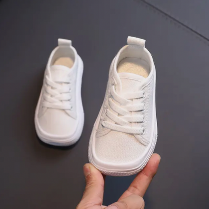 Weiße Kinderschuhe aus Canvas mit Schnürsenkeln, handgehalten, auf dunklem Hintergrund.