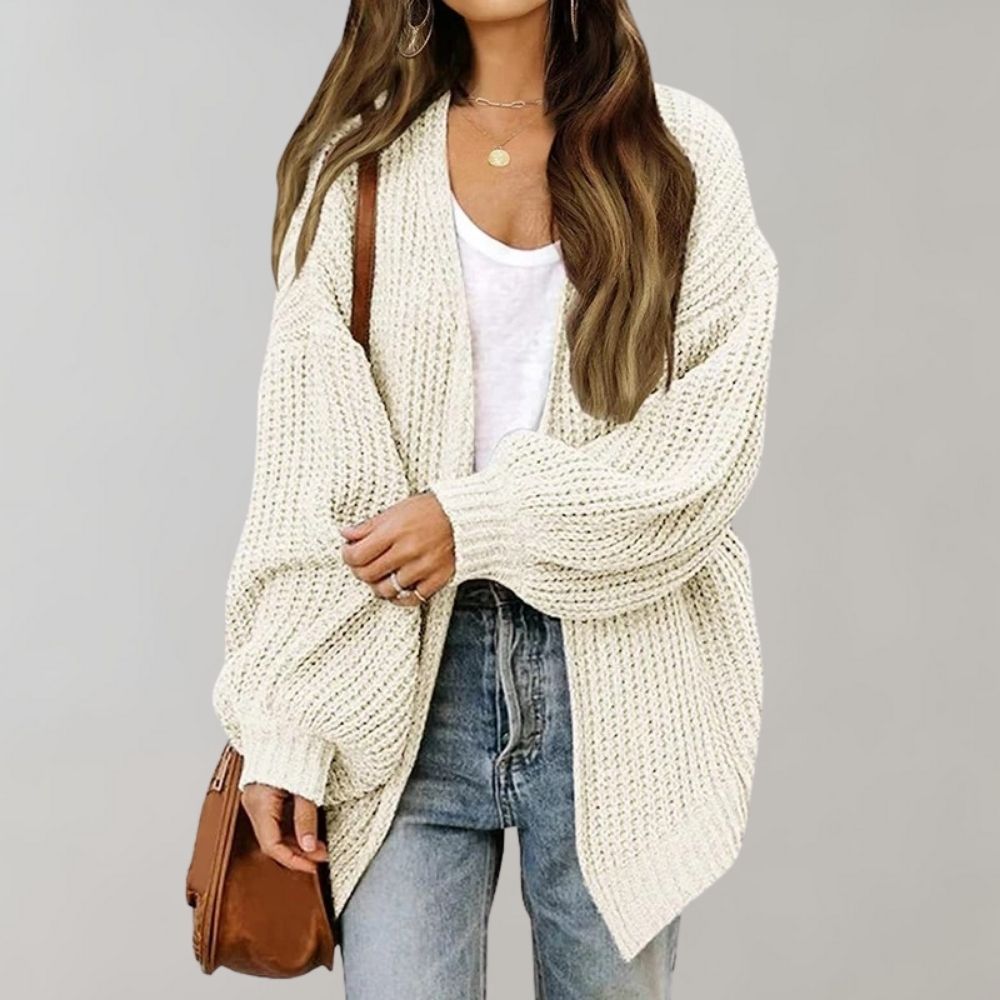 Frau in cremefarbenem Strickcardigan, Jeans und brauner Tasche vor grauem Hintergrund.