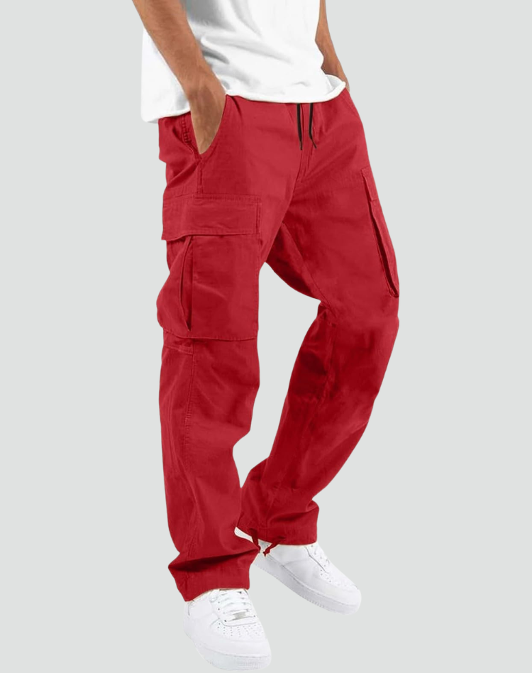 Man bär röda cargobyxor med stora sidfickor och dragsko i midjan, vit t-shirt och vita sneakers, mot grå bakgrund. 10