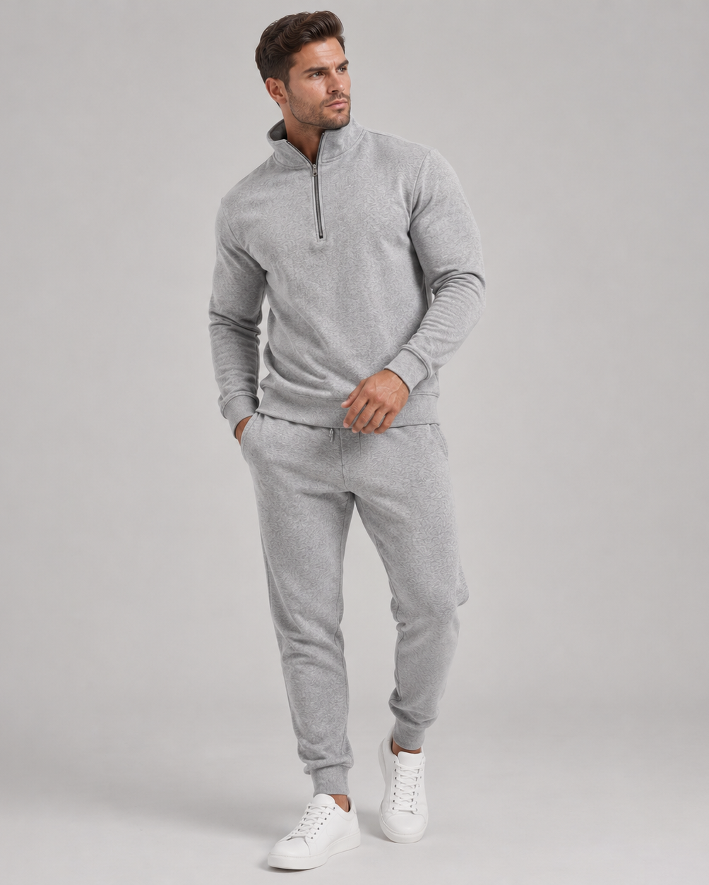 Man i ljusgrått set med halvdragkedja och joggingbyxor, helfigur med vita sneakers mot grå bakgrund 1