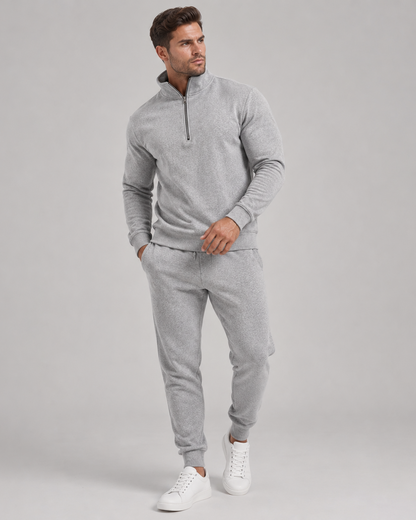 Man i ljusgrått set med halvdragkedja och joggingbyxor, helfigur med vita sneakers mot grå bakgrund 1