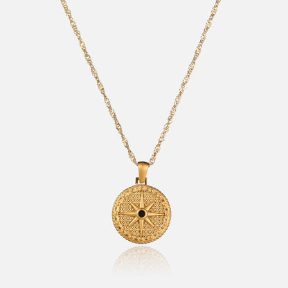 Guldfärgat halsband med rund hängsmycke format som en åttauddig stjärna med en liten svart sten i mitten, sett framifrån mot ljus bakgrund. 1