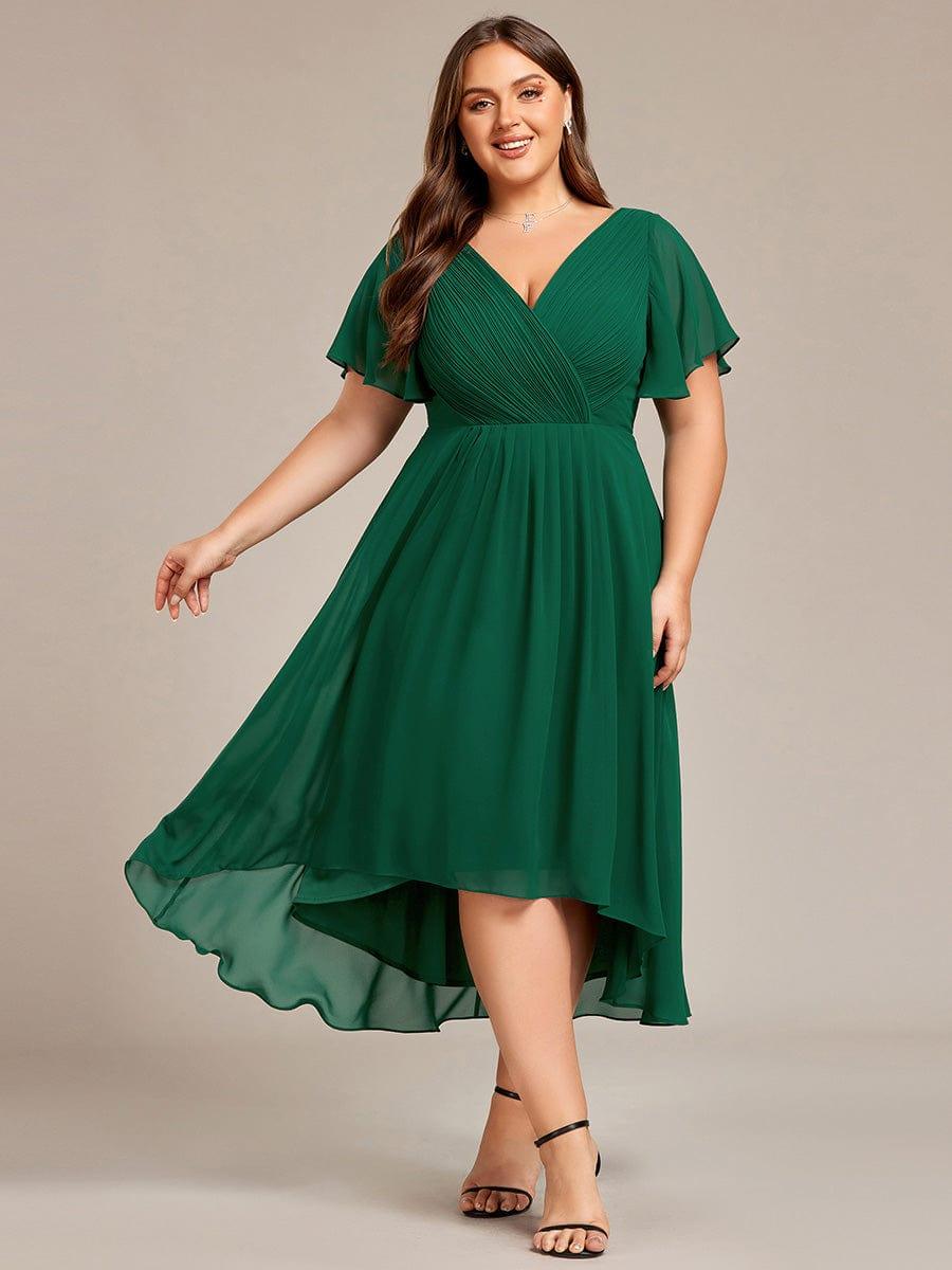 Mörkgrön klänning på plus size-modell som sträcker ut ena armen så att den flödande, asymmetriska kjolen syns, framifrån. 36