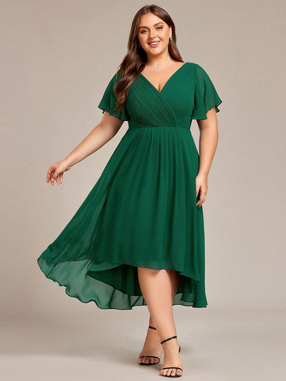 Mörkgrön klänning på plus size-modell som sträcker ut ena armen så att den flödande, asymmetriska kjolen syns, framifrån. 36