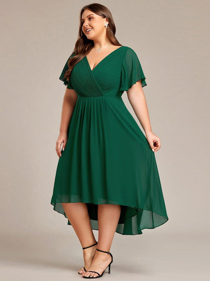 Mörkgrön klänning på plus size-modell snett framifrån, där den högre fronten och längre bakkanten syns tydligt. 39