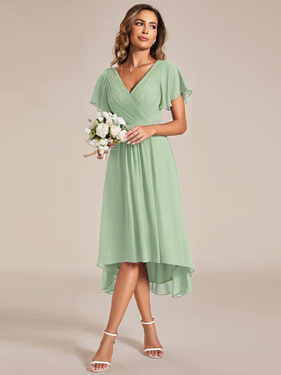 Modell i saltgrön chiffongklänning håller en bukett vita blommor, snett framifrån, med asymmetrisk fåll. 79