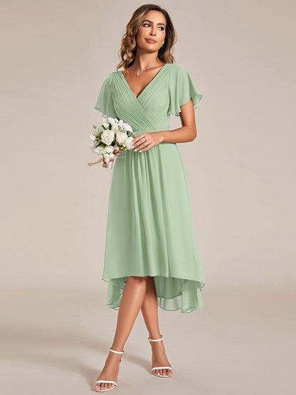 Modell i saltgrön chiffongklänning håller en bukett vita blommor, snett framifrån, med asymmetrisk fåll. 79
