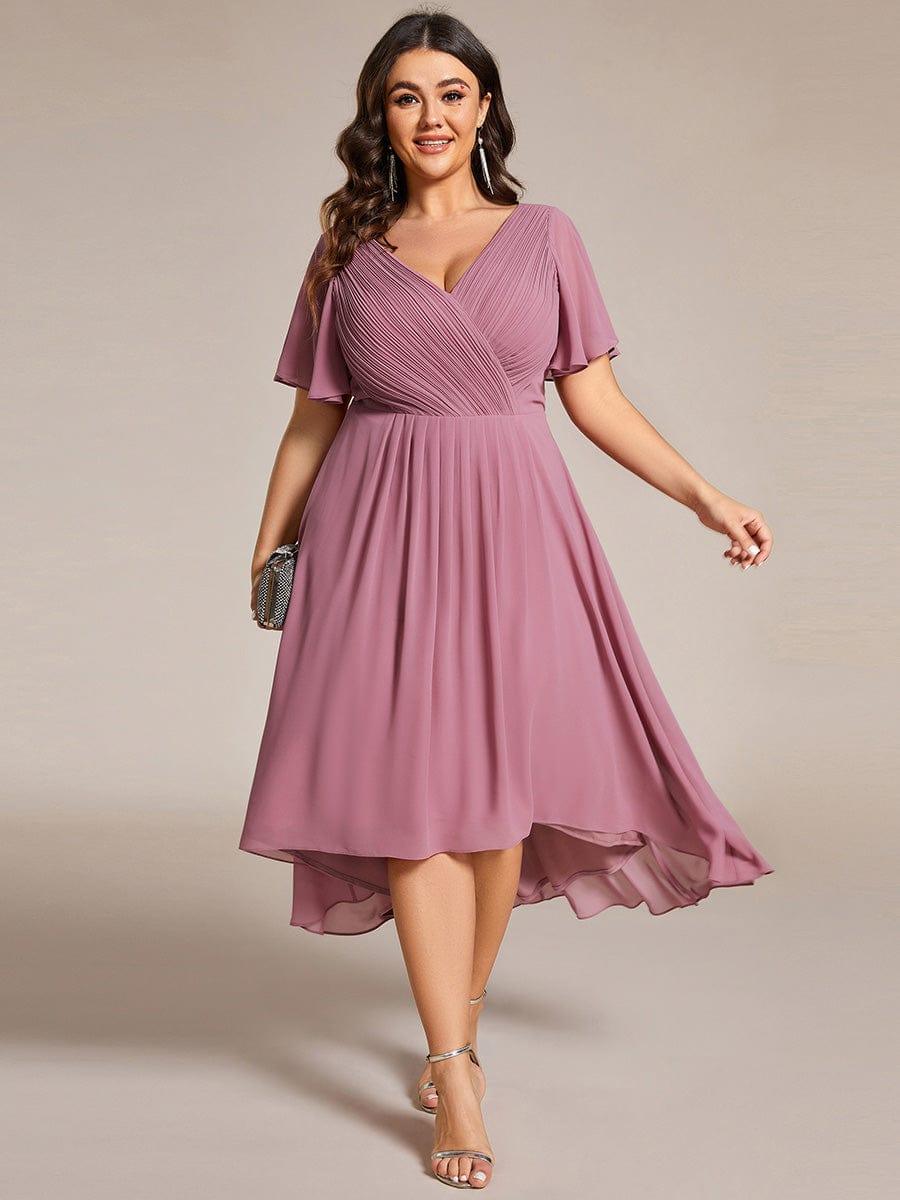 Plus size-modell i dammrosa klänning, framifrån, med V-ringning, korta ärmar, vid kjol och kortare fram, samt silverfärgad clutch. 63
