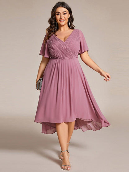 Plus size-modell i dammrosa klänning, framifrån, med V-ringning, korta ärmar, vid kjol och kortare fram, samt silverfärgad clutch. 63