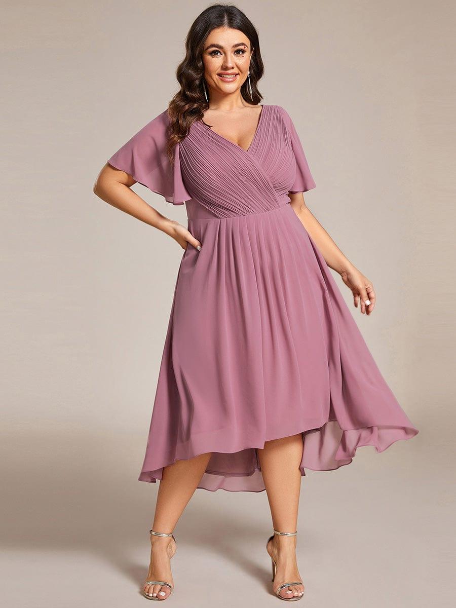 Plus size-modell med handen på höften i dammrosa chiffongklänning med asymmetrisk fållkant och veckad omlottöverdel. 66
