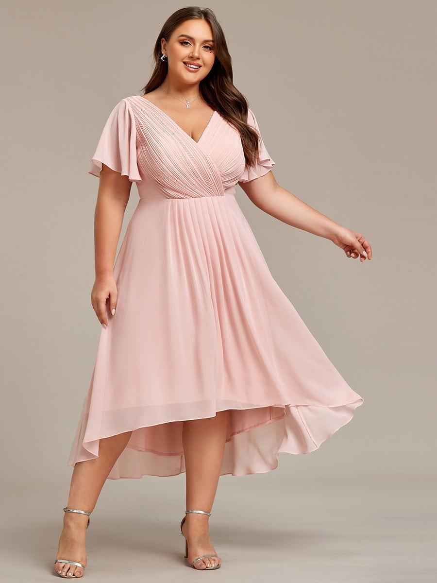 Ljusrosa klänning på leende plus size-modell med ena armen utsträckt, så att den asymmetriska chiffongkjolen böljar. 34