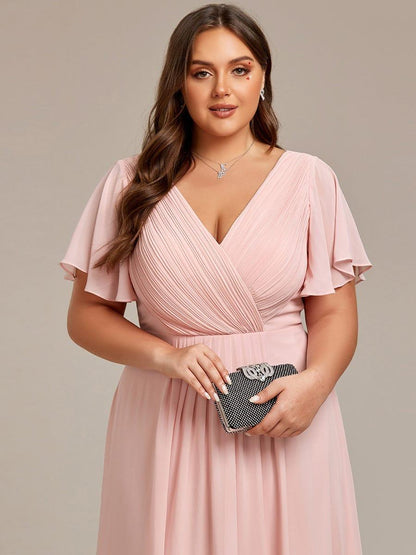 Närbild på ljusrosa klänning med veckat omlott-liv och vida vingärmar, modellen håller en glittrande clutch framför sig. 33