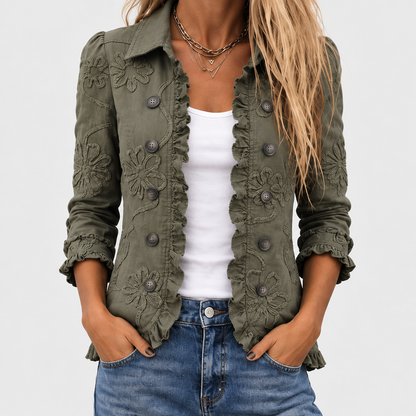 Närbild av den olivgröna jackan på en modell, med broderade blommor, knappar och volangkant längs den öppna framdelen, över vitt linne och jeans. 1