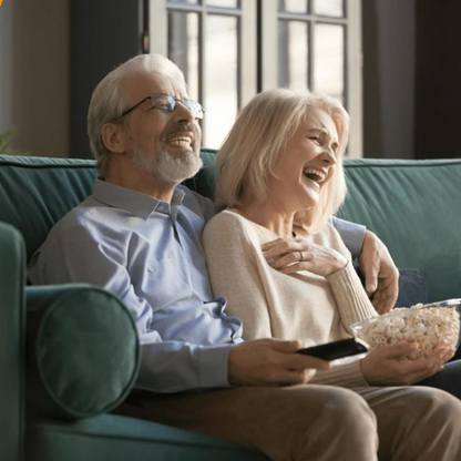 Älteres Paar auf Sofa, lächelnd, mit Popcorn und Fernbedienung, gemütliches Wohnzimmer.