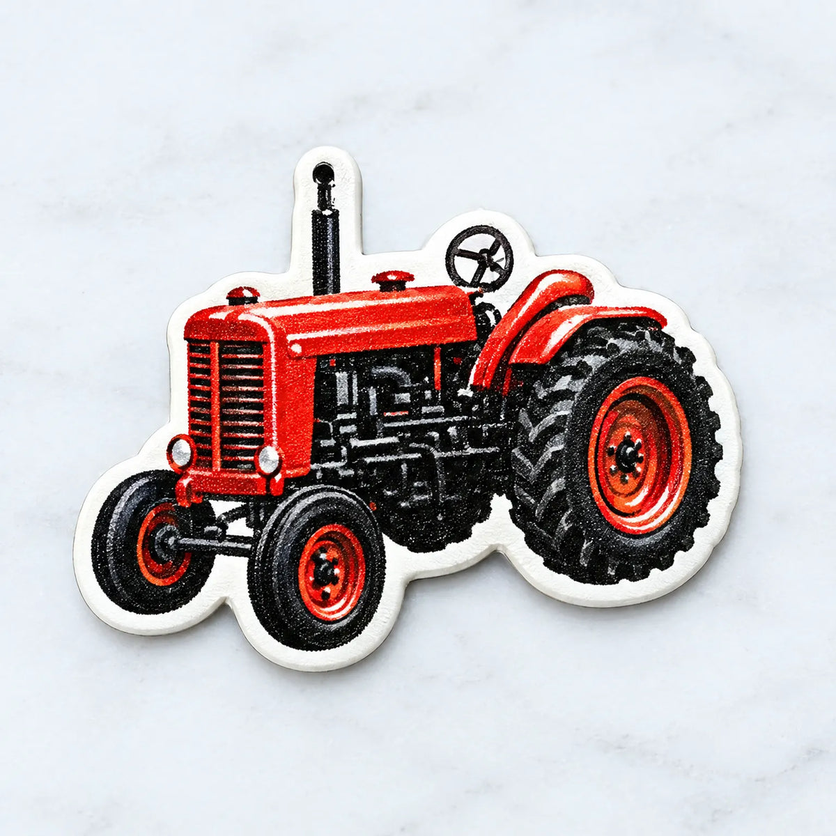 Närbild på en röd traktor i form av en dekoration med hål upptill för upphängning, liggande på en ljus marmoryta. 3