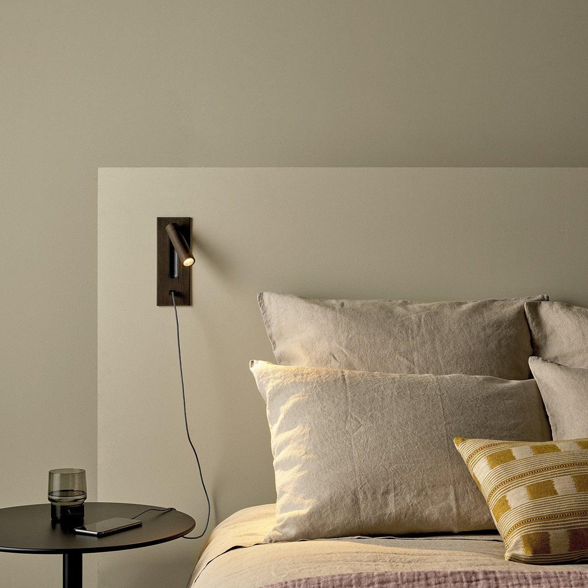 Bronsfärgad vägglampa med USB-kabel kopplad till en mobil på ett litet svart sängbord 36