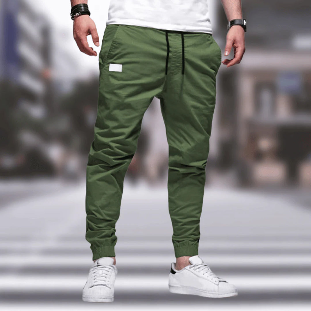 Mann trägt grüne Jogginghose, weißes T-Shirt, weiße Sneaker, urbaner Hintergrund.