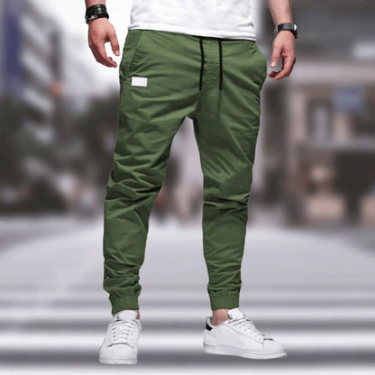 Mann trägt grüne Jogginghose, weißes T-Shirt, weiße Sneaker, urbaner Hintergrund.