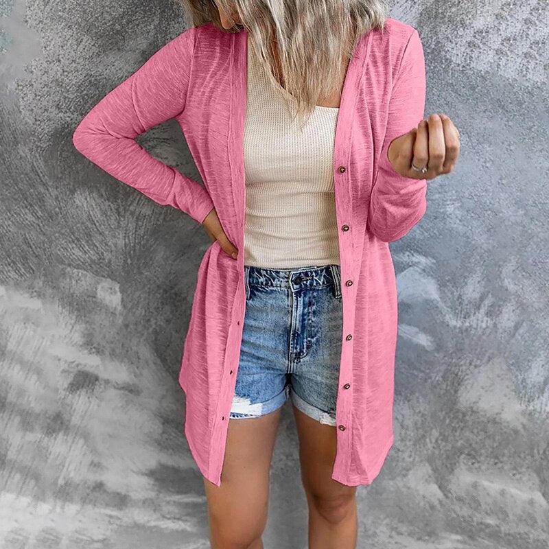 Frau in rosa Strickjacke, weißem Top und Jeansshorts vor grauem Hintergrund.