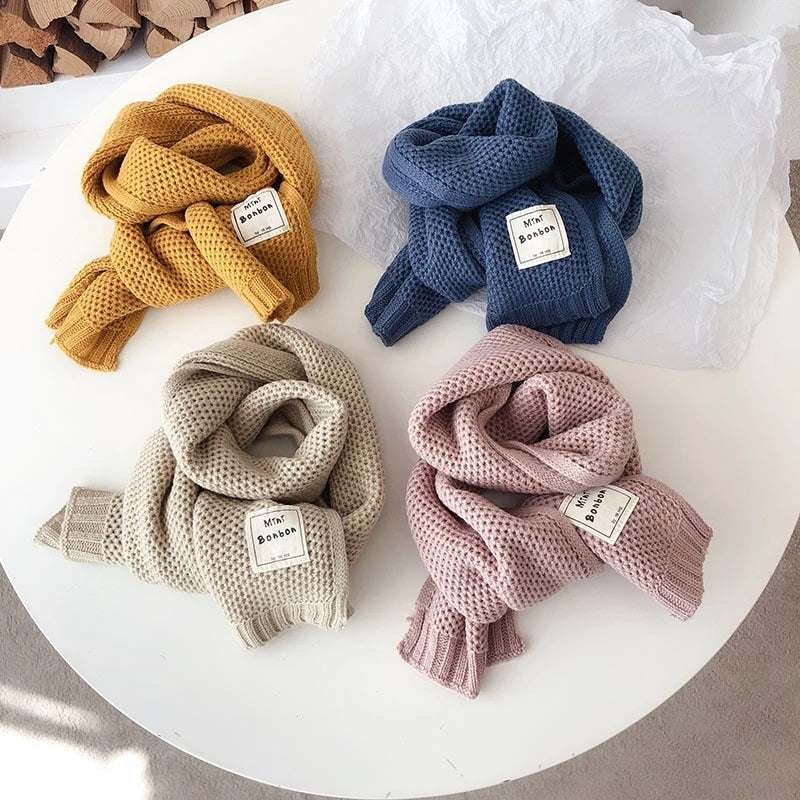 Vier gestrickte Schals in Gelb, Blau, Beige und Rosa auf weißem Tisch, weiches Material, modisch.