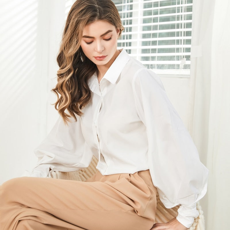 Frau in weißer Bluse und beiger Hose, sitzt auf Stuhl, elegantes Büro-Outfit.
